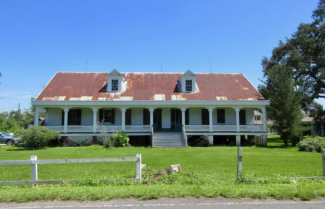 White raised plantation house with a long front gallery and rusted red metal roof behind a lawn