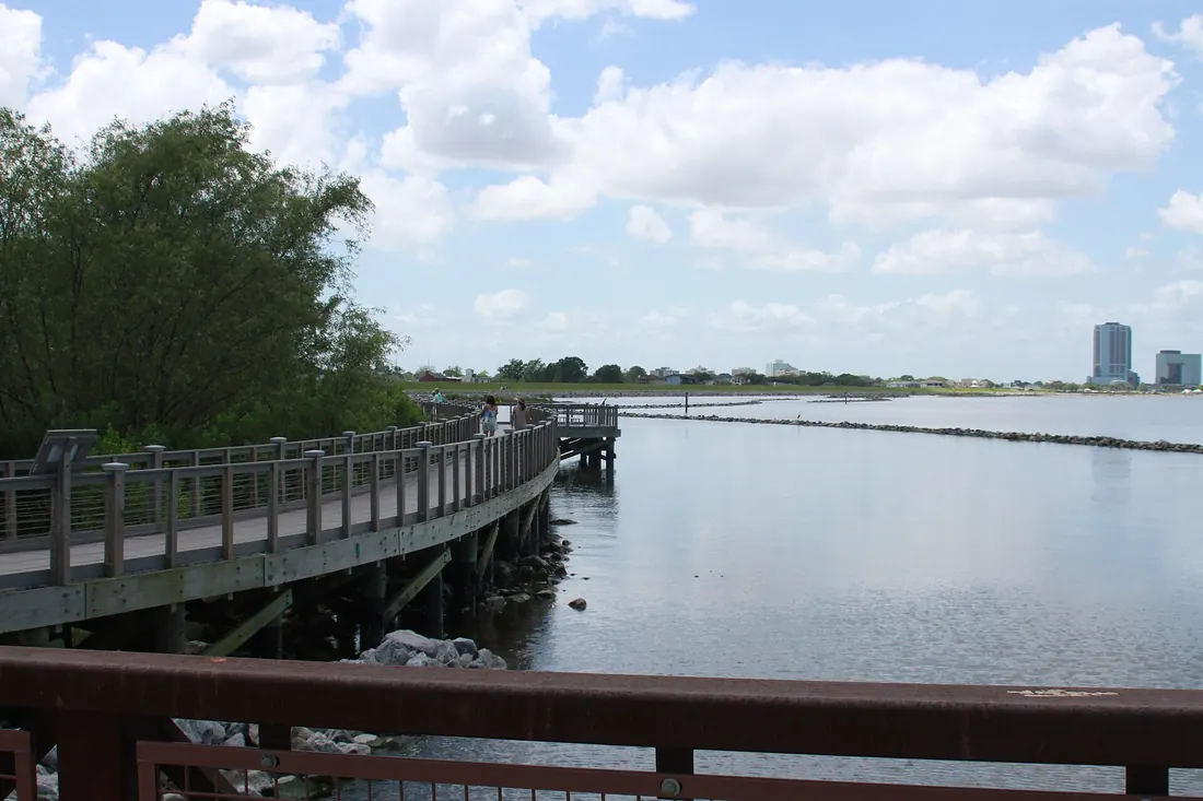 Wooden boardwalk curving along calm Lake Pontchartrain water with trees, breakwaters and distant high-rise buildings