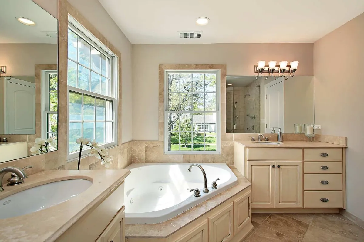 Elegant bathroom with a large corner bathtub under two windows, beige cabinets, and a marble counter