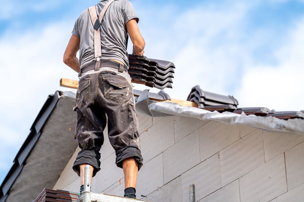 A construction worker stands on scaffolding, placing roof tiles on a house under construction beneath a blue sky—an everyday scene in roof repair New Orleans projects.