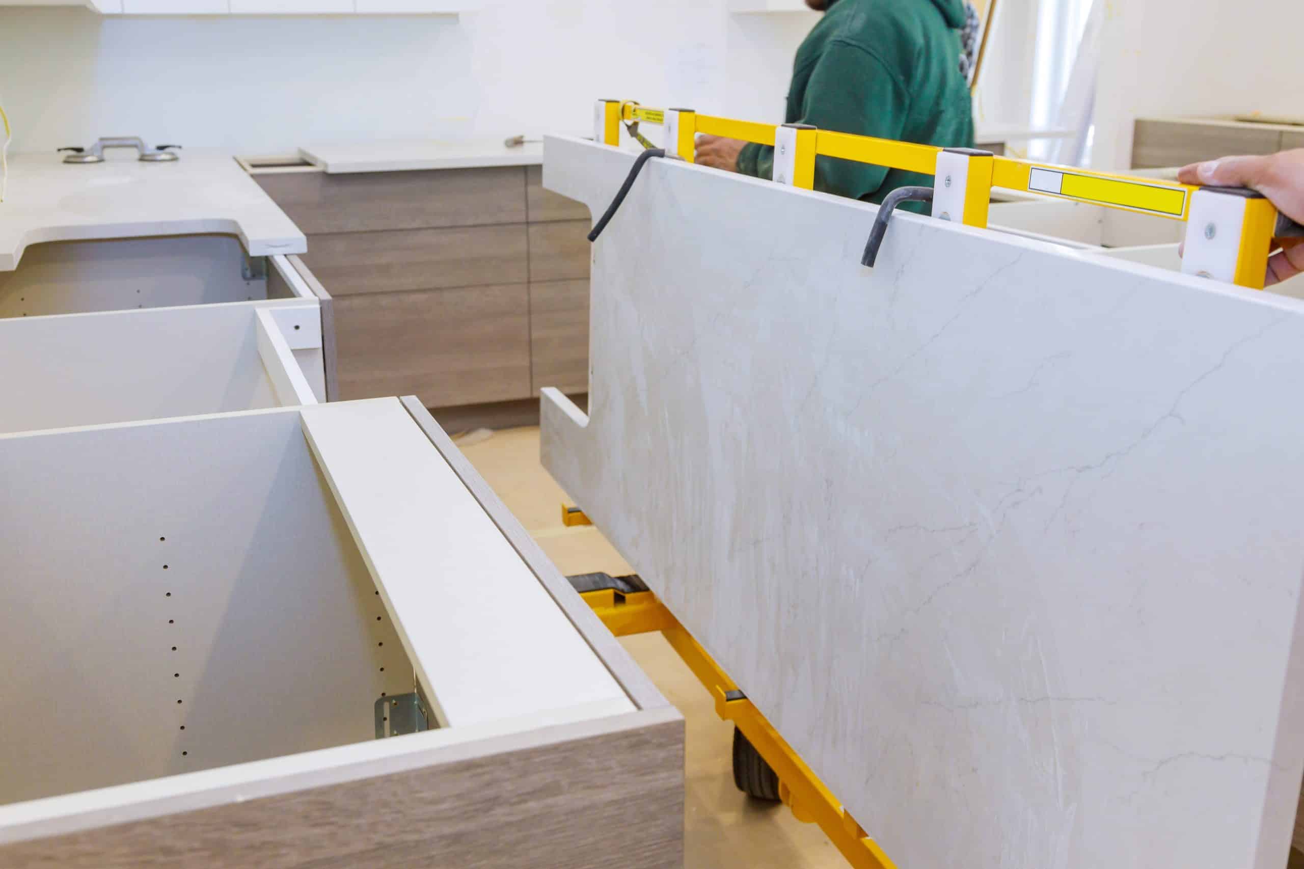 Two people use a yellow lifting tool to position the kitchen countertop to be installed onto lower cabinets in a partially finished kitchen.
