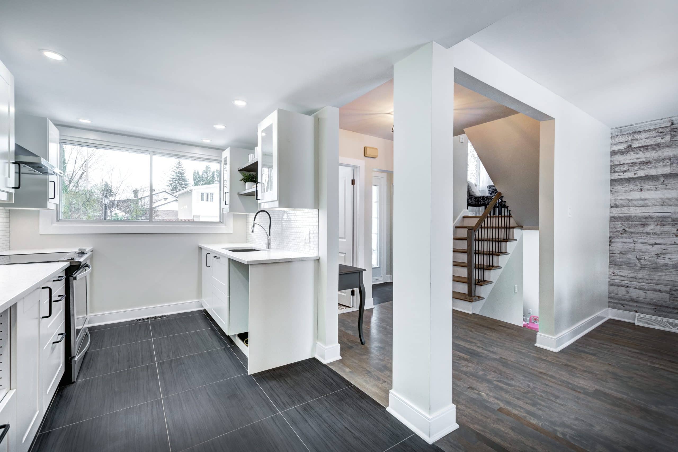 Modern kitchen with white cabinets, black countertops, and a large window showcases the quality of this whole-home renovation. An open entryway leads to a staircase with wood steps and a gray textured accent wall.