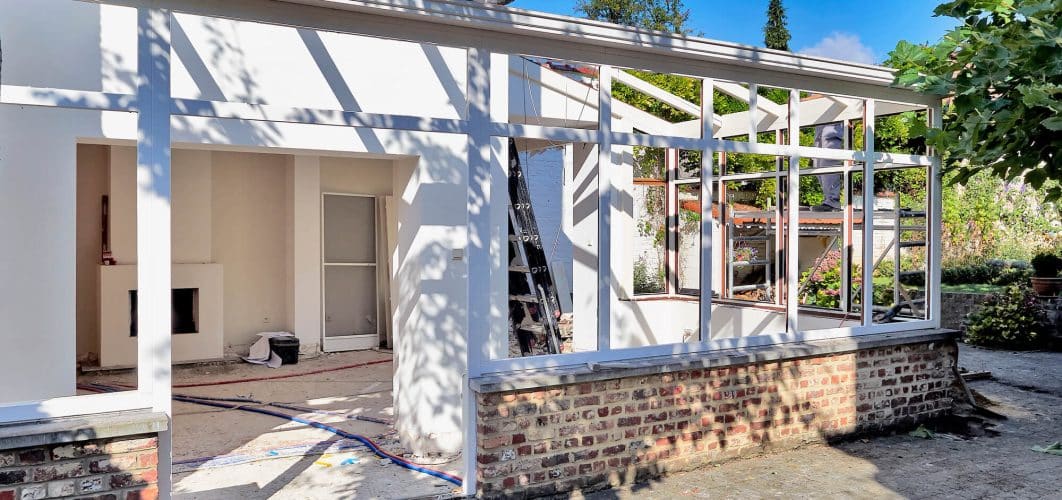 A partially constructed home addition to a brick house with exposed framework and unfinished walls, with construction materials visible inside and outside.