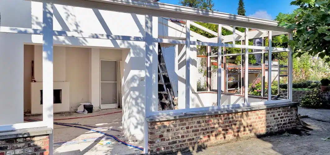 A partially constructed home addition to a brick house with exposed framework and unfinished walls, with construction materials visible inside and outside.