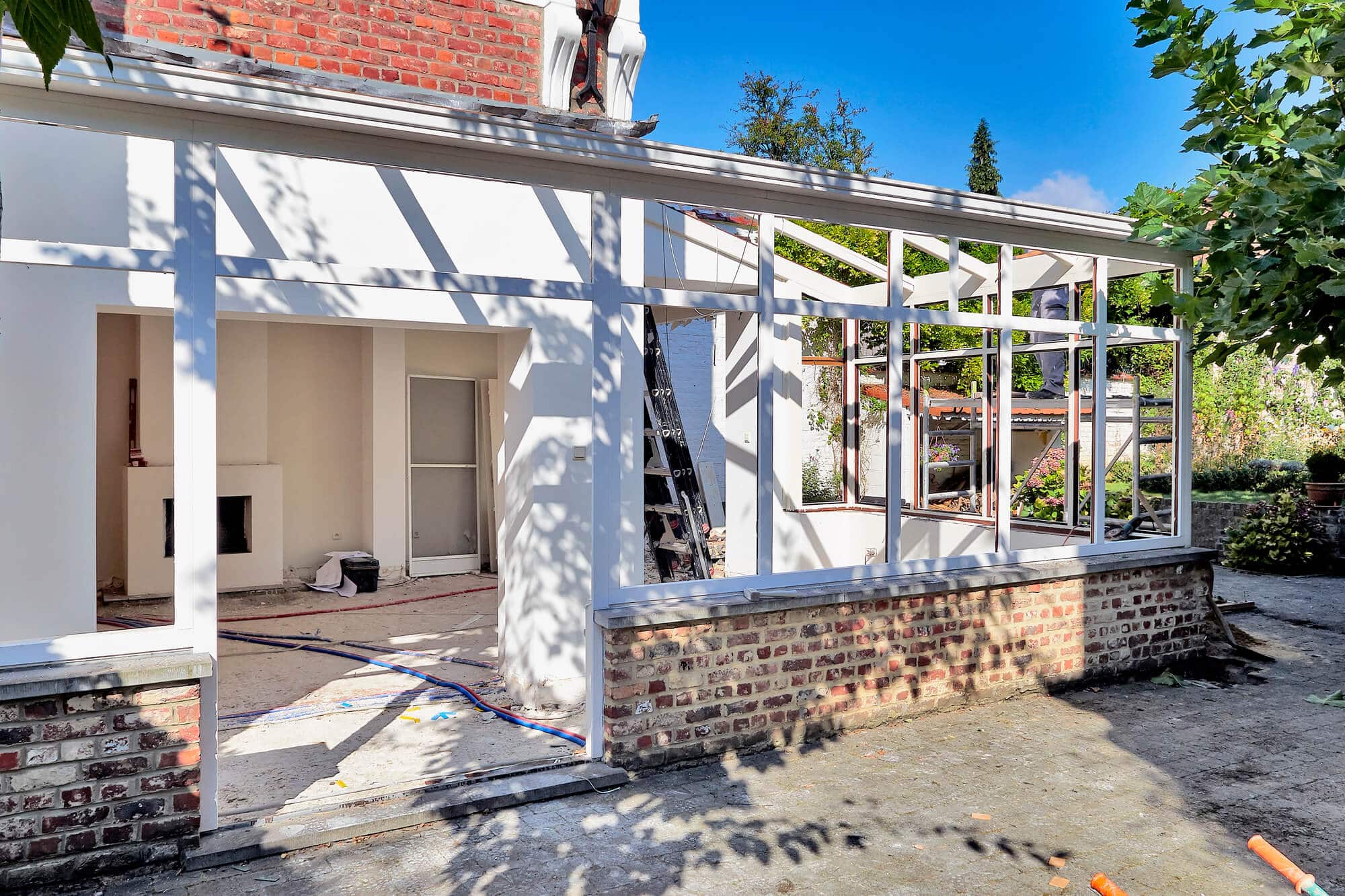 A partially constructed home addition to a brick house with exposed framework and unfinished walls, with construction materials visible inside and outside.