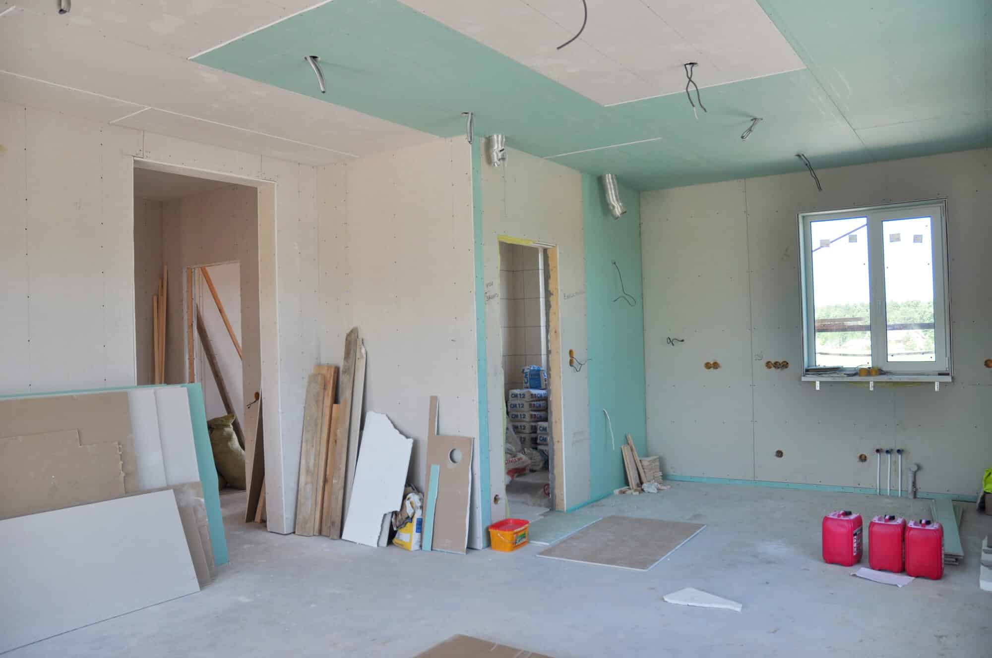 Room under construction for room expansion, featuring unfinished walls, exposed wires, drywall sheets, construction materials on the floor, and a window on the right wall.