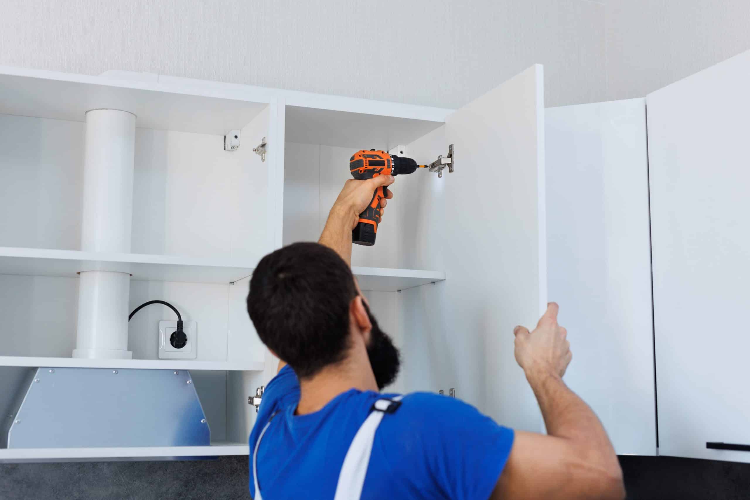 A man uses a power drill to install or adjust the door on a white kitchen cabinet.