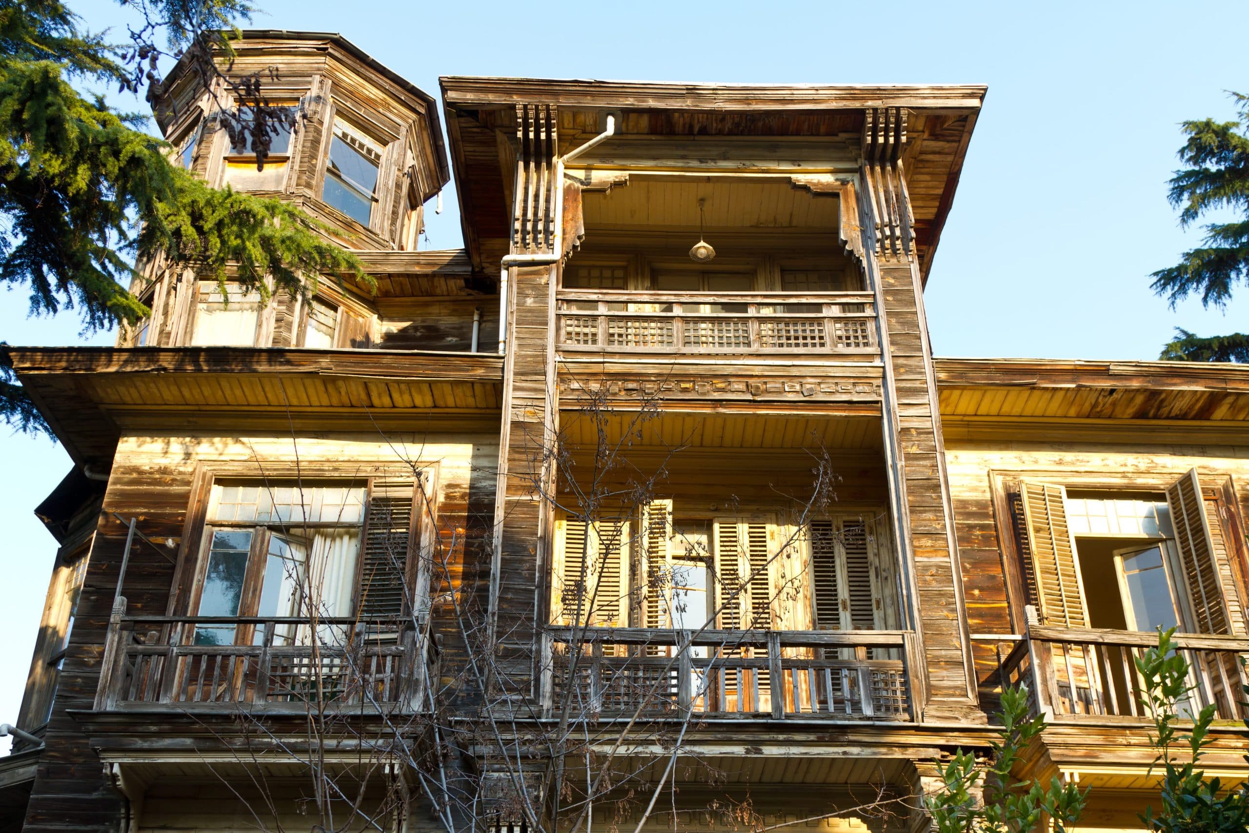 A weathered, two-story wooden house with a small tower, several balconies, and shuttered windows, surrounded by trees. 