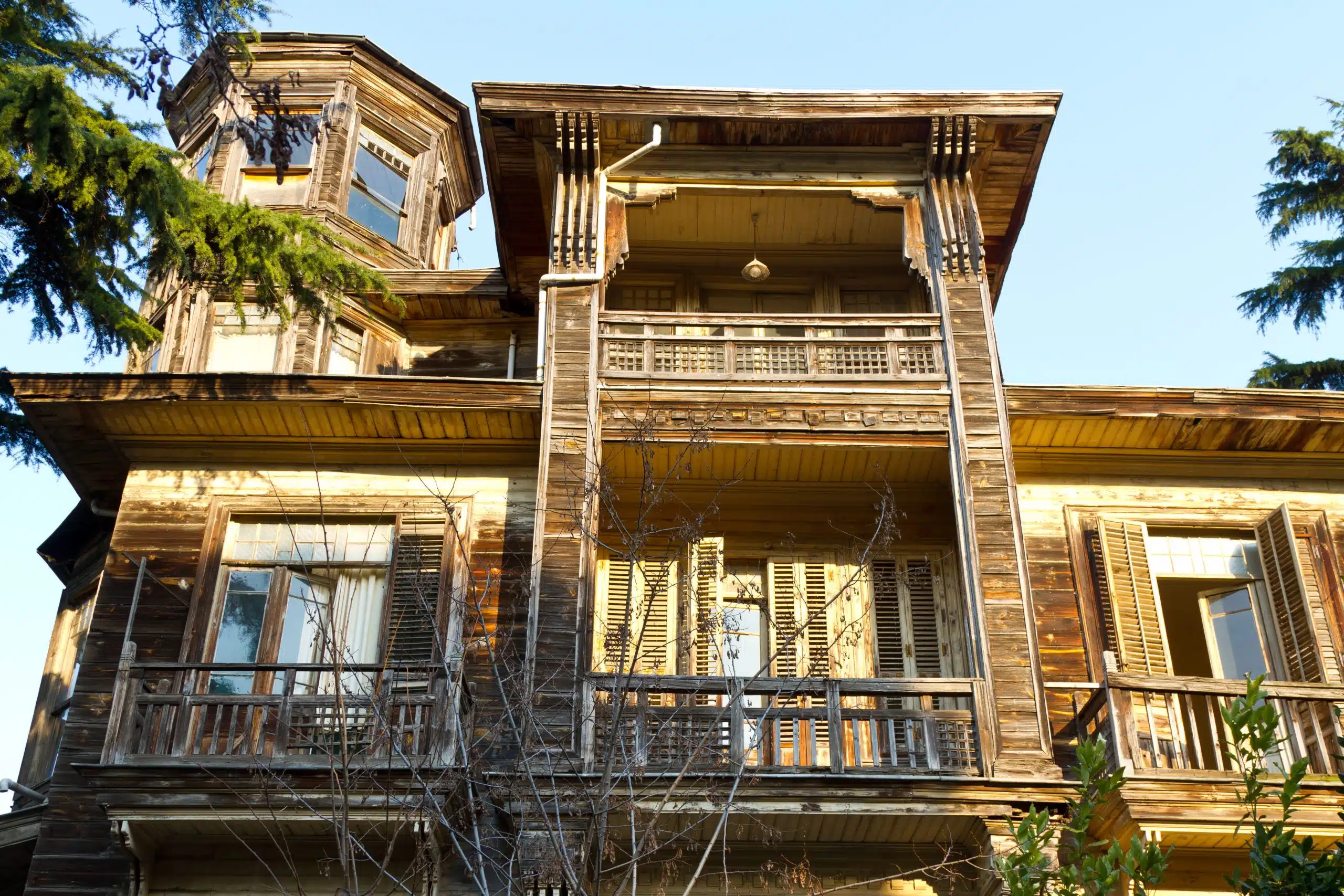 A weathered, two-story wooden house with a small tower, several balconies, and shuttered windows, surrounded by trees. 