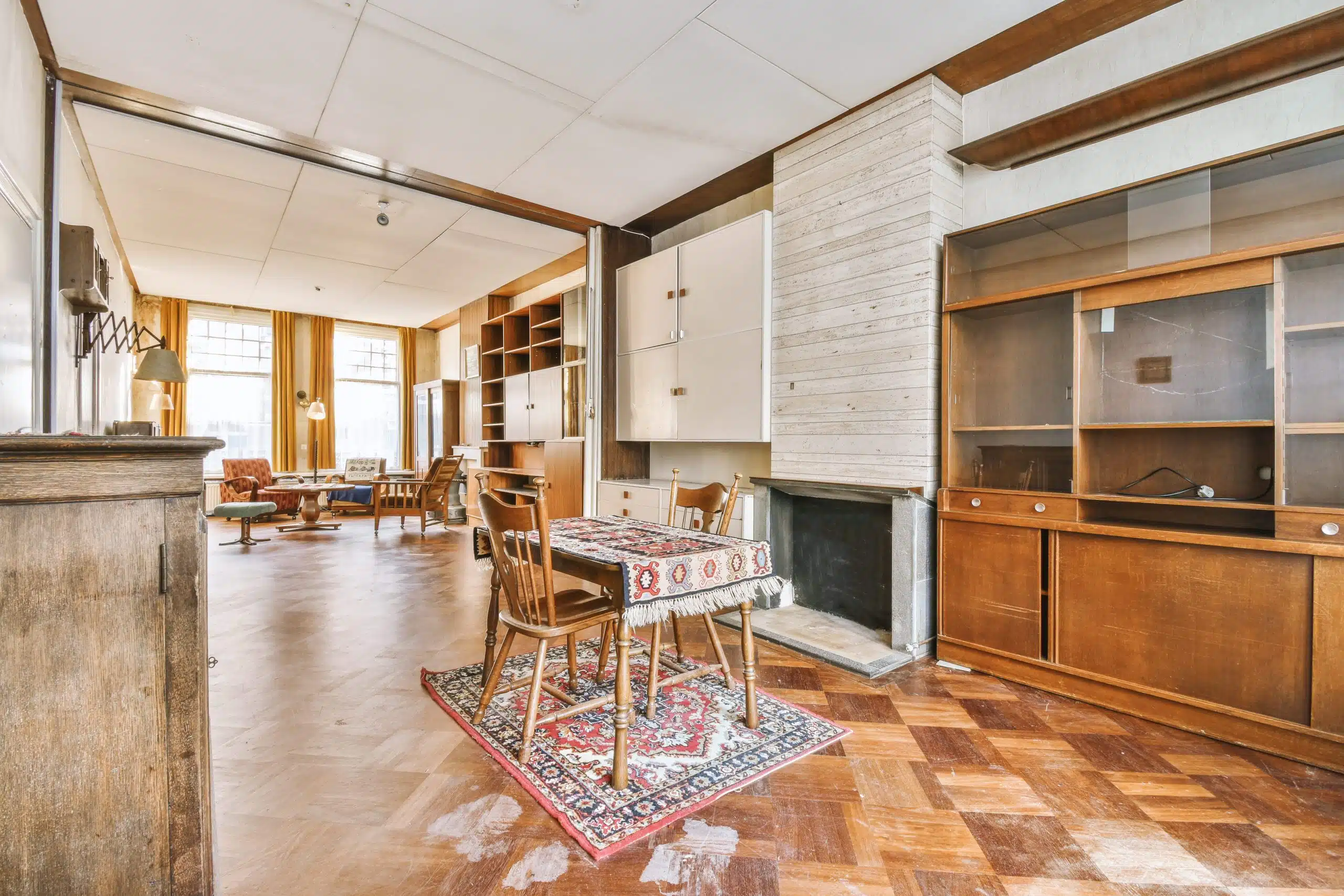 A vintage living and dining room with wooden furniture, parquet flooring, a small dining table on a rug, built-in cabinets, and large windows with yellow curtains reflects an outdated home interior charm.