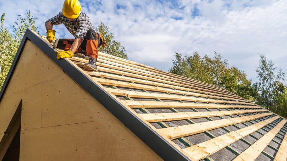 A construction worker wearing a yellow hard hat installs roof boards on a house under construction, representing the skilled roof repair New Orleans relies on, set against a blue sky with clouds.