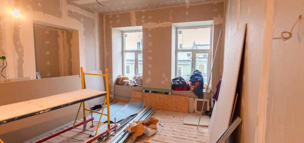 Room under renovation with new drywall hung and taped on the walls and ceiling and a spare sheet leaning against the wall