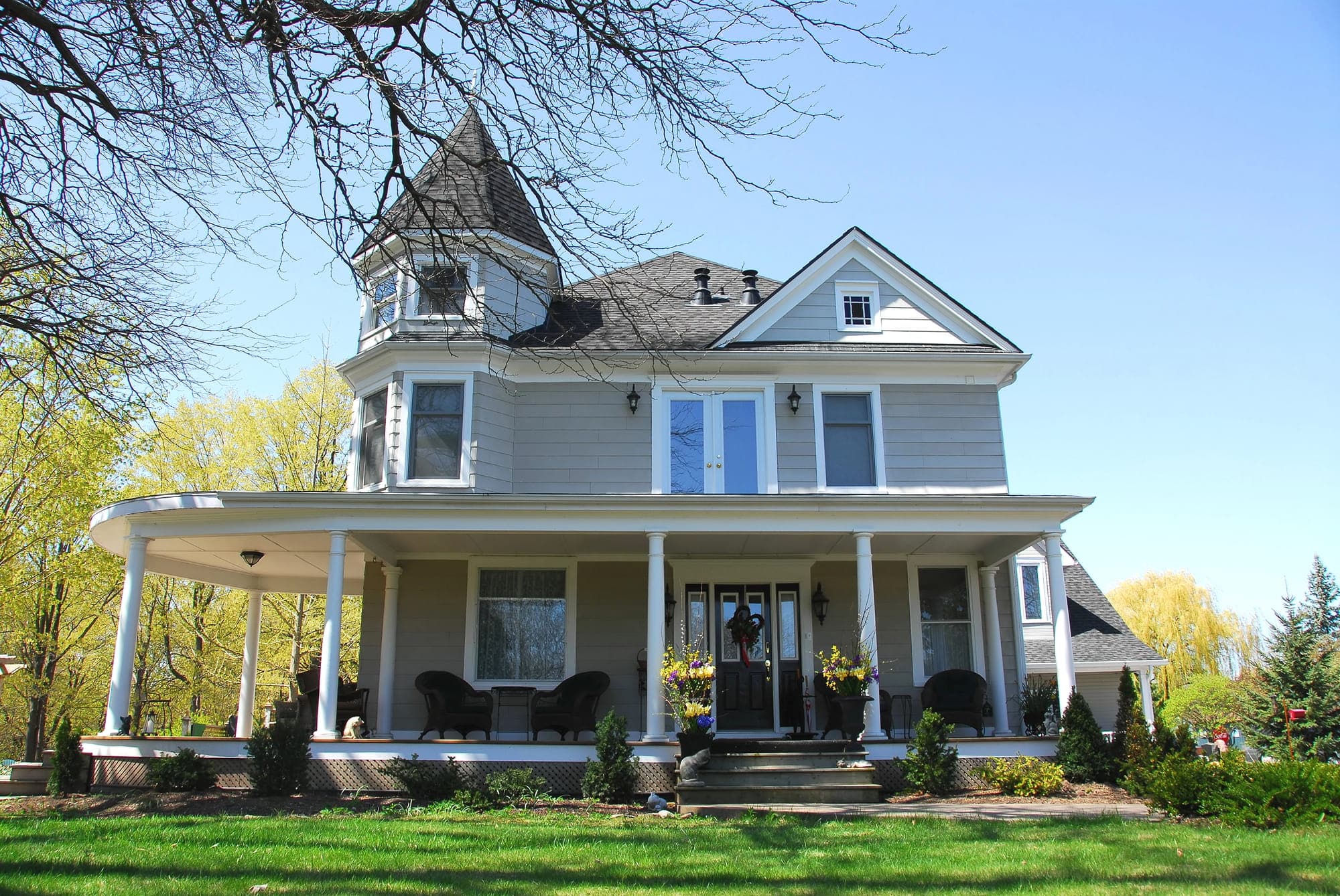 Charming Victorian style house with a turret, wraparound porch, and multiple windows, surrounded by lush shrubs and trees on a sunny day.