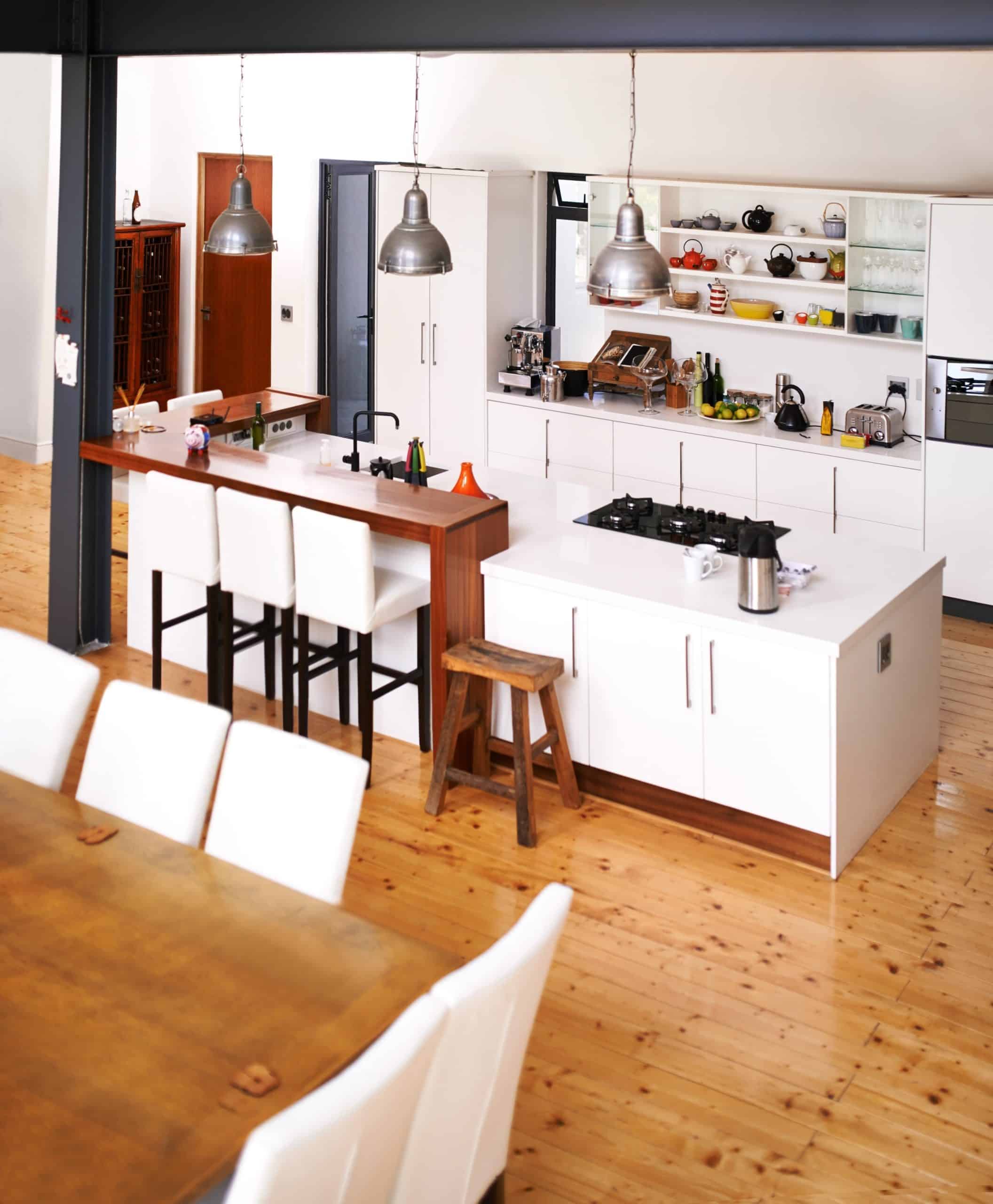 Modern open-plan kitchen with white cabinets, a central island featuring a gas cooktop, pendant lights, bar stools, and a wooden dining table with white chairs on a polished kitchen floor.