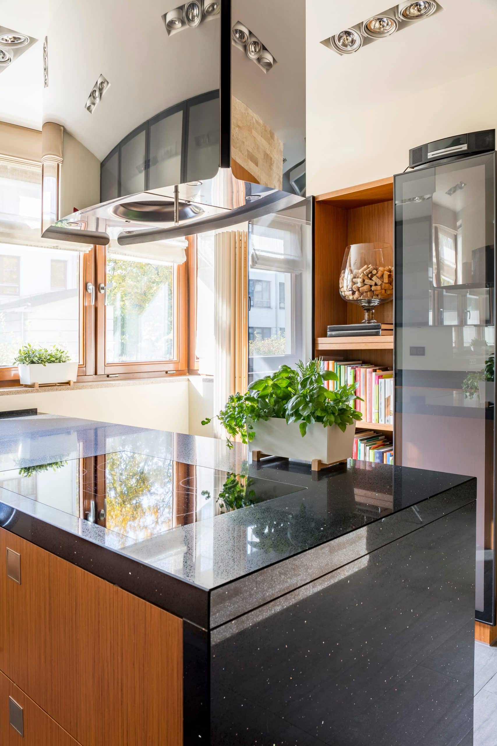 featuring a sleek countertop, electric cooktop, wooden cabinets, potted herbs, bookshelves, large windows, and a stainless steel range hood.