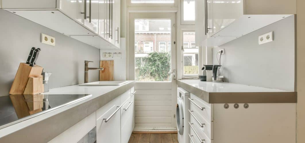 Modern space saving kitchen with white cabinets, sleek countertops, a stovetop, knife block, coffee maker, and washing machine, featuring a glass door that leads to an outdoor area.
