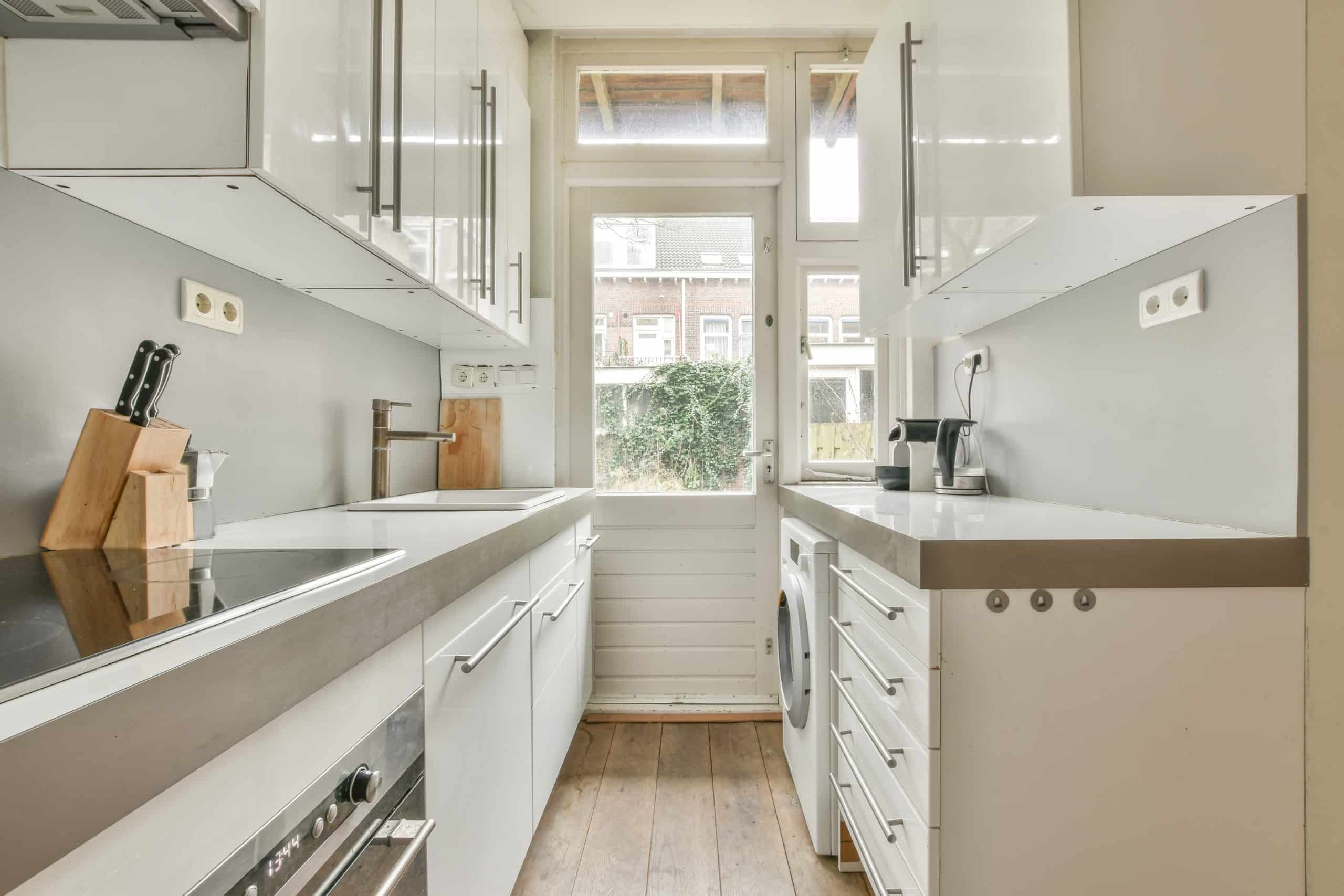 Modern space saving kitchen with white cabinets, sleek countertops, a stovetop, knife block, coffee maker, and washing machine, featuring a glass door that leads to an outdoor area.