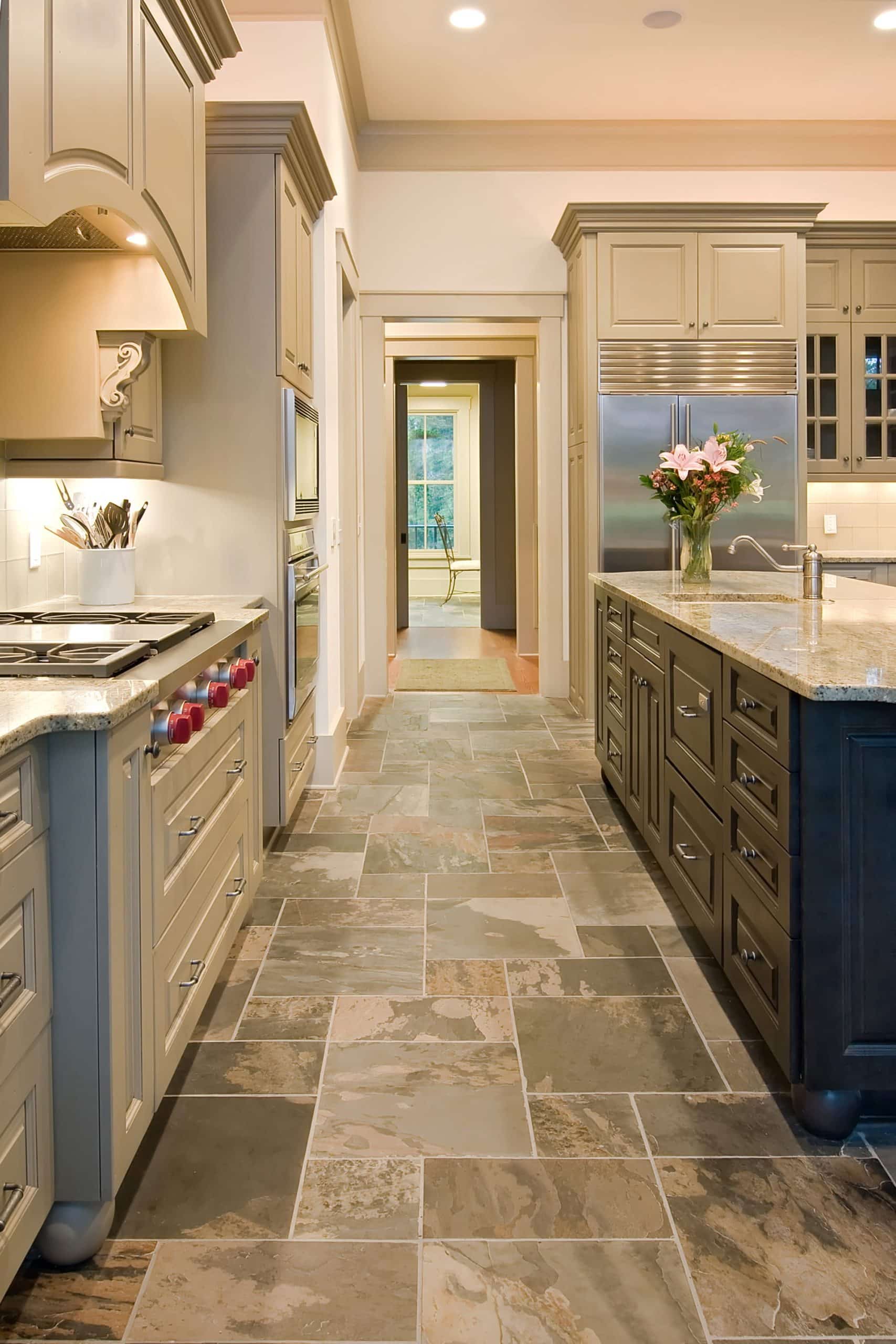 Modern kitchen with beige cabinetry, stainless steel appliances, kitchen floor featuring stone tile, a central island with drawers, and a vase of flowers on the counter. Hallway leads to another room.
